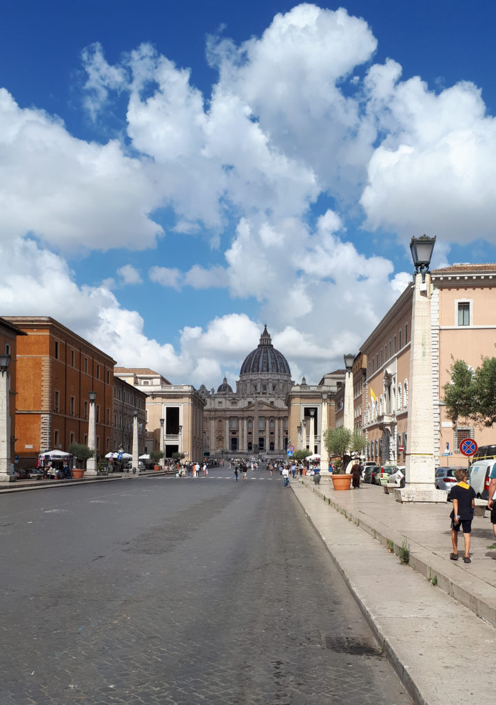 Grande avenue bordé de palais à la façade ocre pour celui de gauche et rose pour celui de droite. Basilique Saint Pierre au fond.