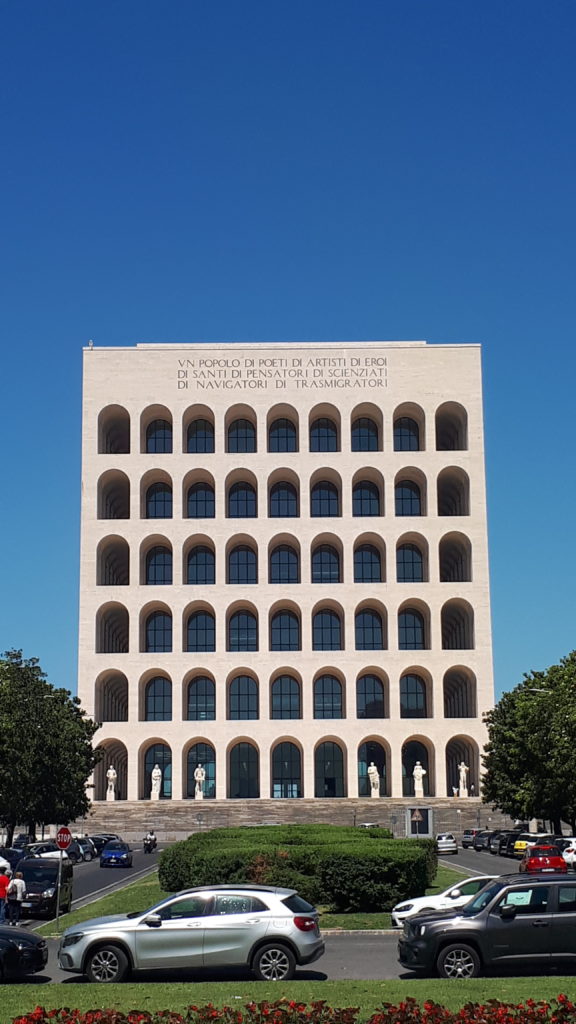 Bâtiment carré, avec une façade composé d'arches (6 en hauteur, 9 de large). Des statues au pied du bâtiment.