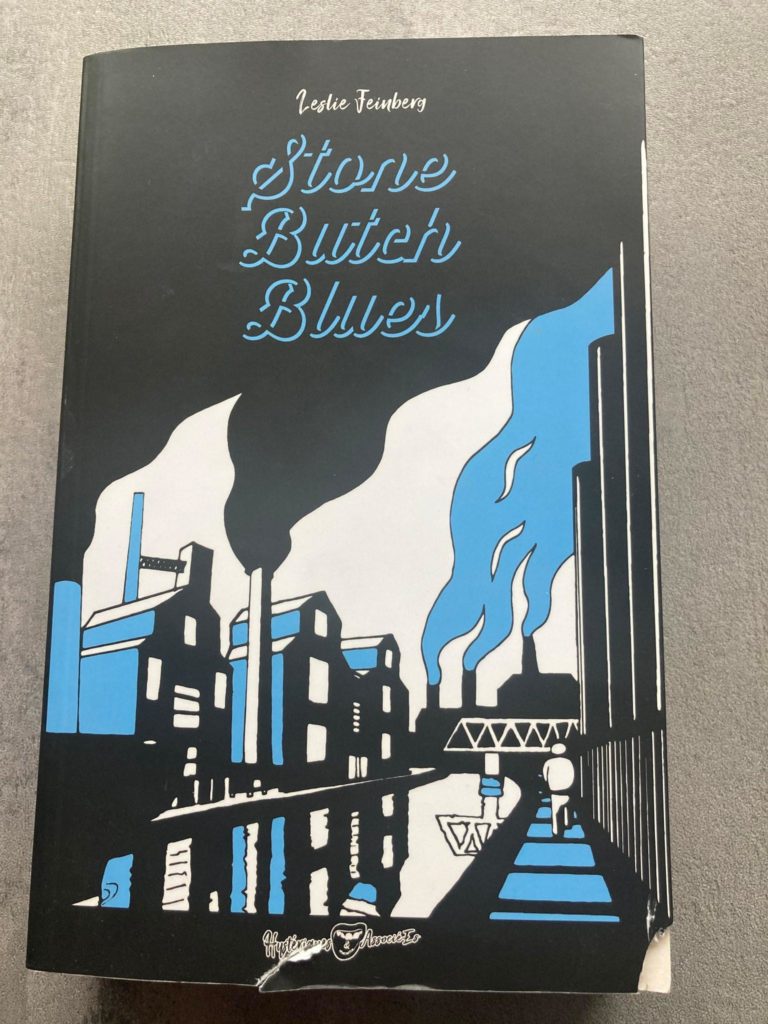 Couverture du livre, en noir, bleu et blanc. Des usines crachent de la fumée. Des silhouettent à leur pied marchent.