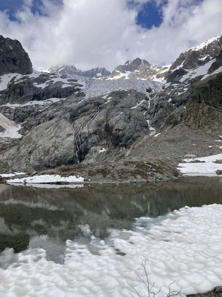 Photo. Un glacier blanc-gris, fissuré de milliers d'entailles, sur une montagne aux roches grises.
Au pied de la montagne, un  lac aux bords enneigés qui reflète le glacier. Le ciel bleu est nuageux.