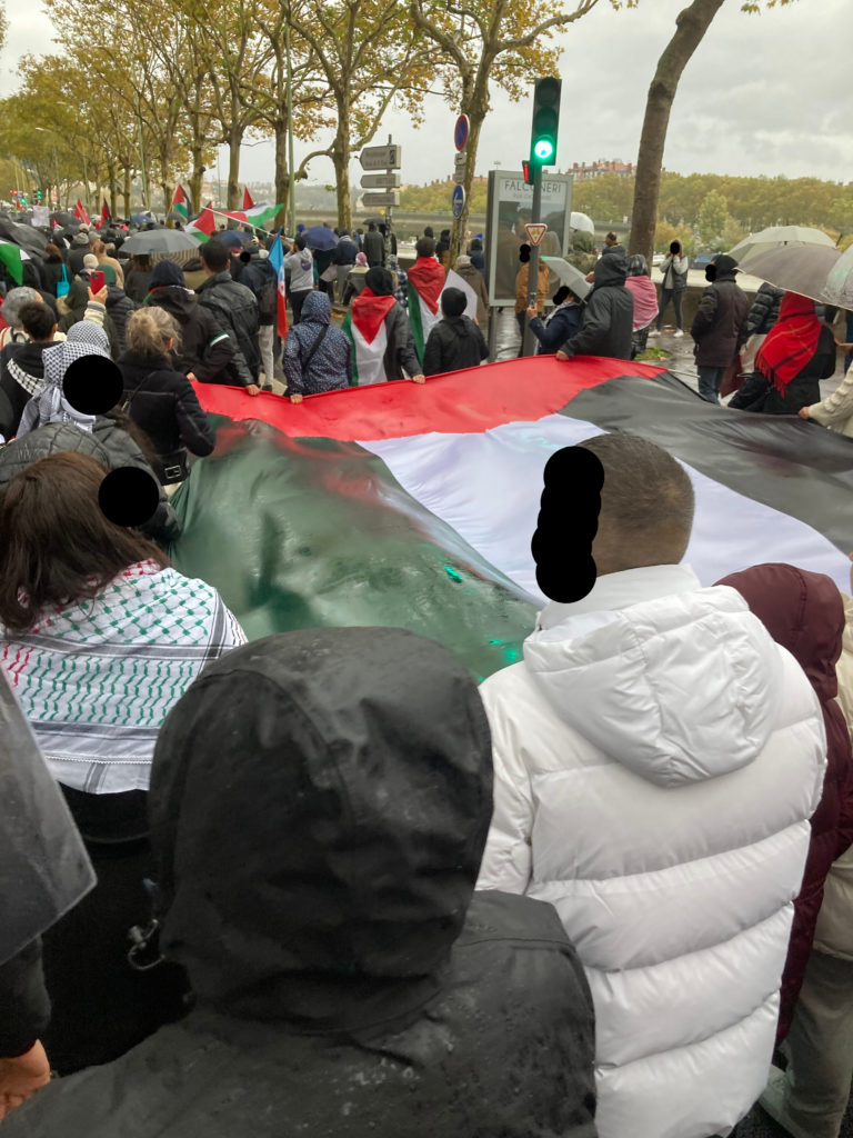 Photo d'une manifestation sous la pluie.
En premier plan, des gens tiennent un énorme drapeau palestinien.
De nombreux drapeaux palestiniens sont tenus par la foule.