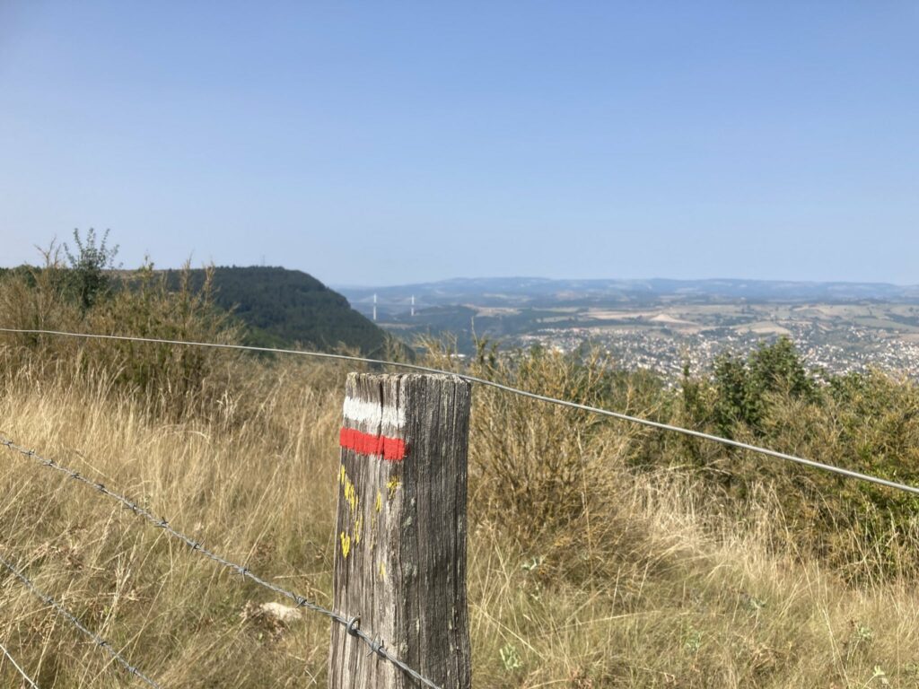 Photo d'un poteau de clôture avec les marques rouges et blanches indiquant un parcours de grande randonnée.
En fond, on devine le viaduc de Millau en arrière plan