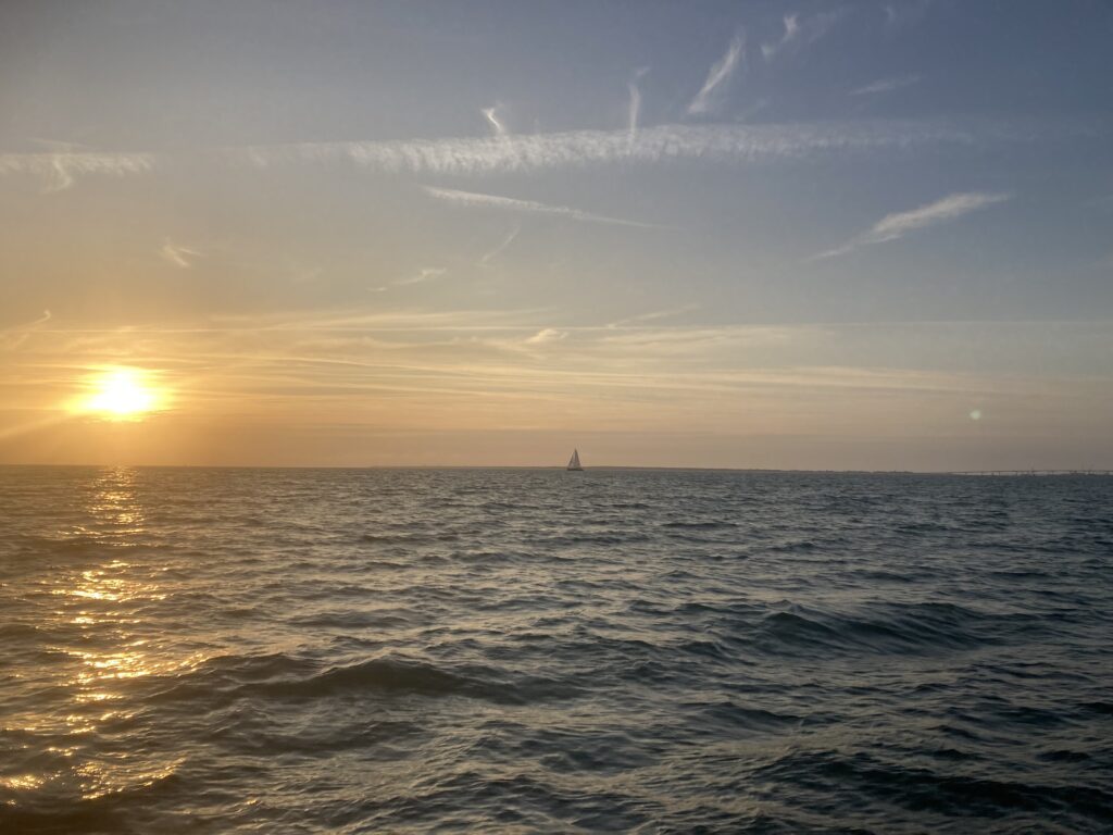 Photo de l'océan au coucher de soleil.
au premier plan, des vagues agitées.
Loin à l'horizon, un voilier blanc.
On aperçoit la lune à gauche de l'horizon.